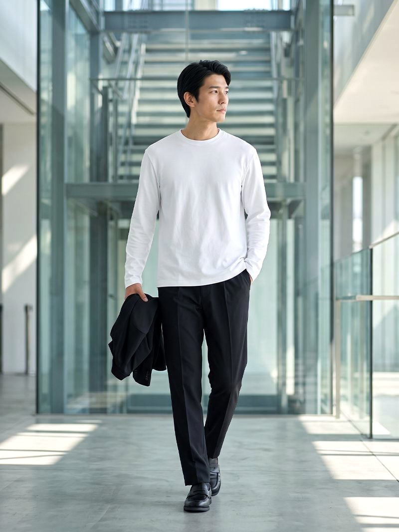 ブラックのパンツに白のロングスリーブT。ジャケットを手に持ってガラス張りの階段前を歩く