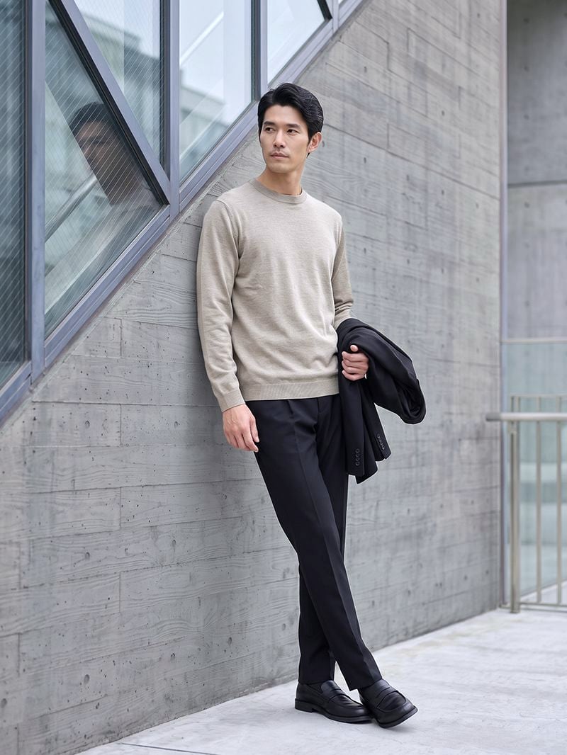 ブラックのパンツにベージュのニット。ジャケットを腕にかけてコンクリートの壁にもたれる