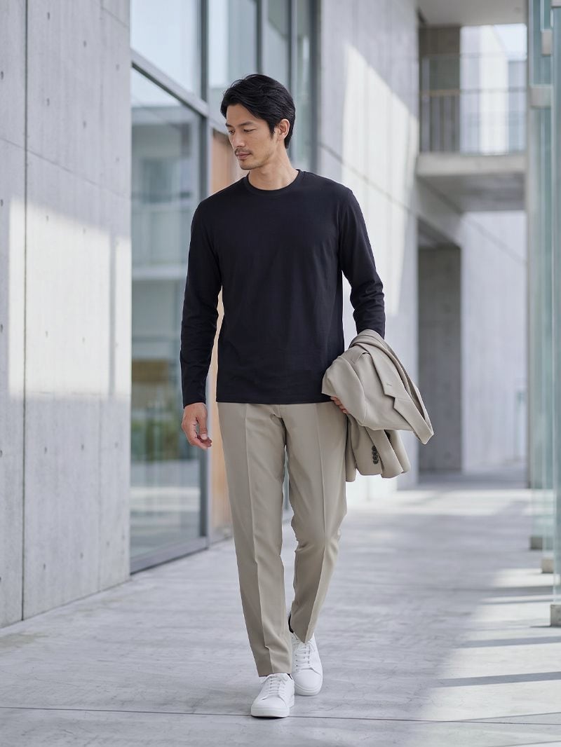 ブラックのCODEロングスリーブTシャツにベージュのパンツ。ベージュのジャケットを手に持って歩く男性