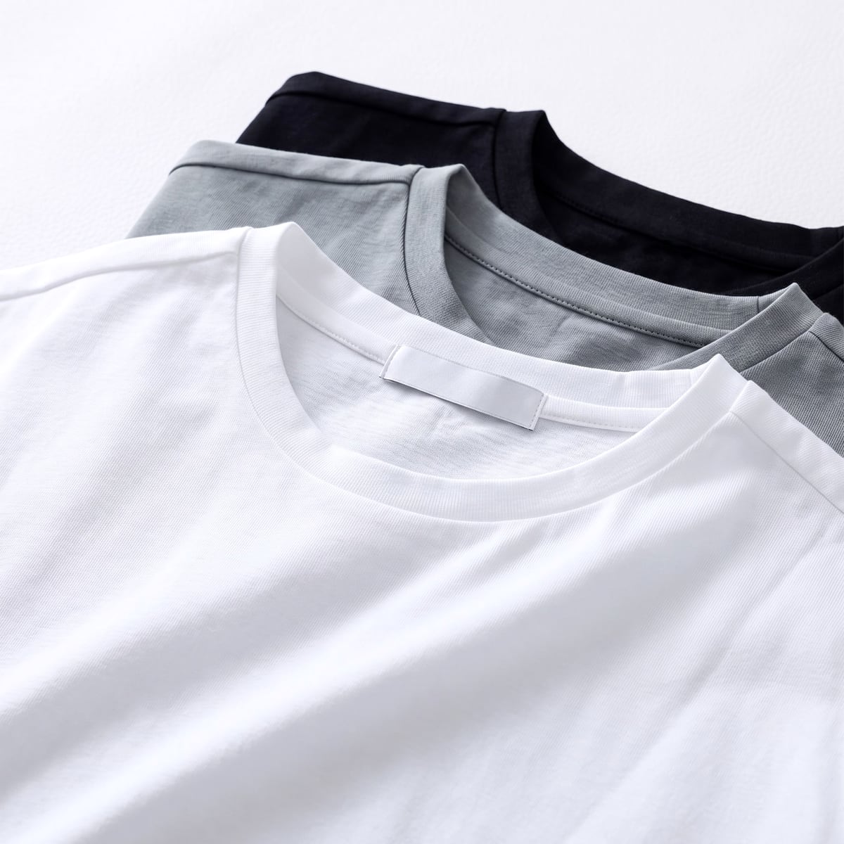 白・グレー・黒の半袖Tシャツを重ねた平置き。なめらかな表面の寄り